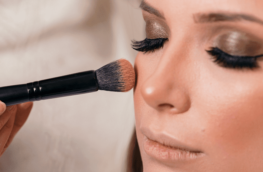 Makeup brush applying blush to woman's cheek, highlighting her glamorous eye makeup.