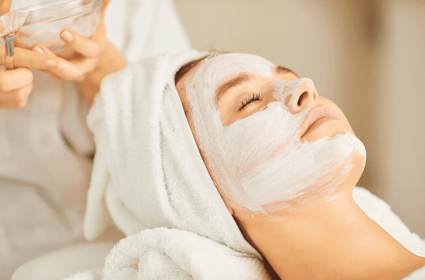 Person with a white face mask relaxing at a spa, head wrapped in a towel, receiving treatment.
