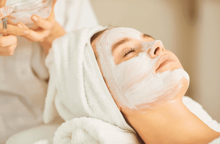 Person with a white face mask relaxing at a spa, head wrapped in a towel, receiving treatment.